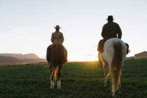 Back view of man and woman riding horses against sunset sky on ranch